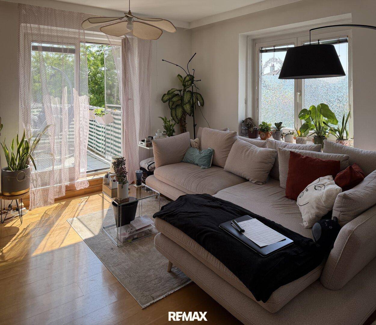 Gemütliches Wohnzimmer mit großem Sofa, vielen Pflanzen und Zugang zu einem Balkon.