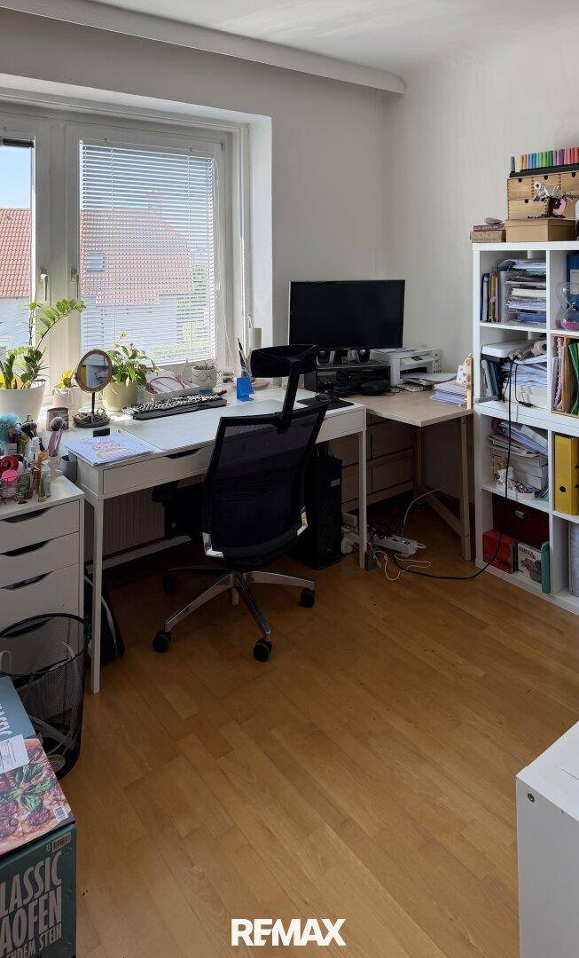 Arbeitszimmer mit Schreibtisch, Bürostuhl, Bücherregal und Fenster mit Blick ins Grüne.