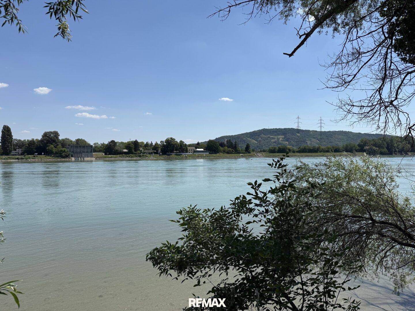 Weitläufiger Blick über einen Fluss mit bewaldeten Hügeln im Hintergrund unter blauem Himmel.