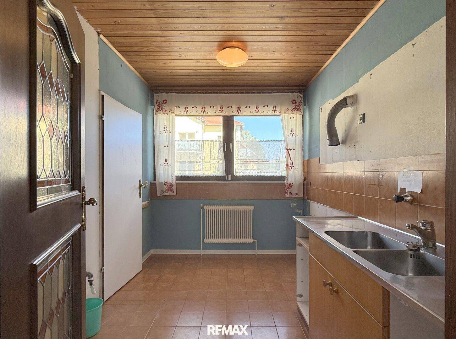 Ältere Küche mit Holzdecke, Fliesenboden und Fensterblick ins Freie.