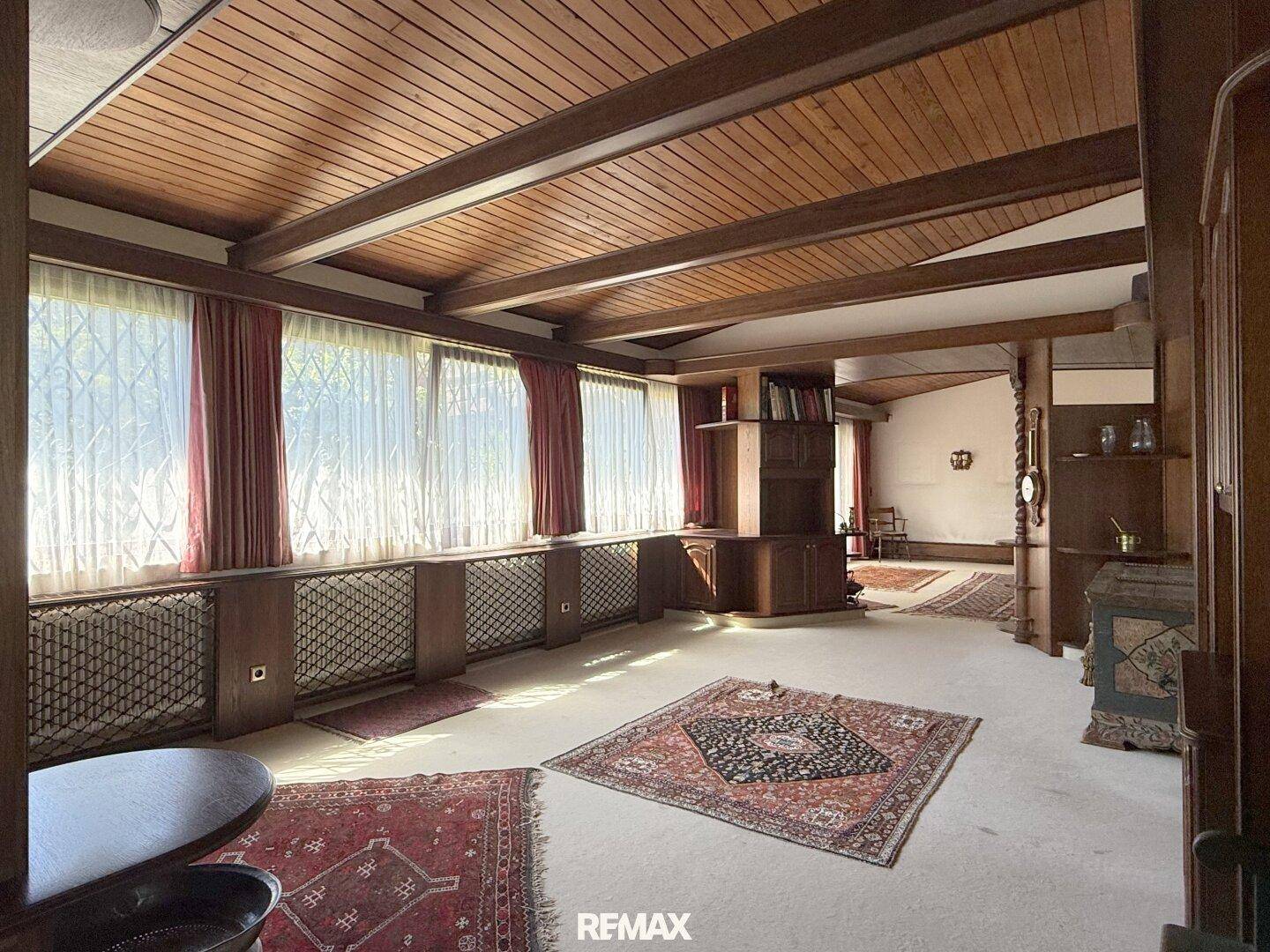 Geräumiges Wohnzimmer mit Holzbalkendecke, Teppichboden und großen Fenstern.