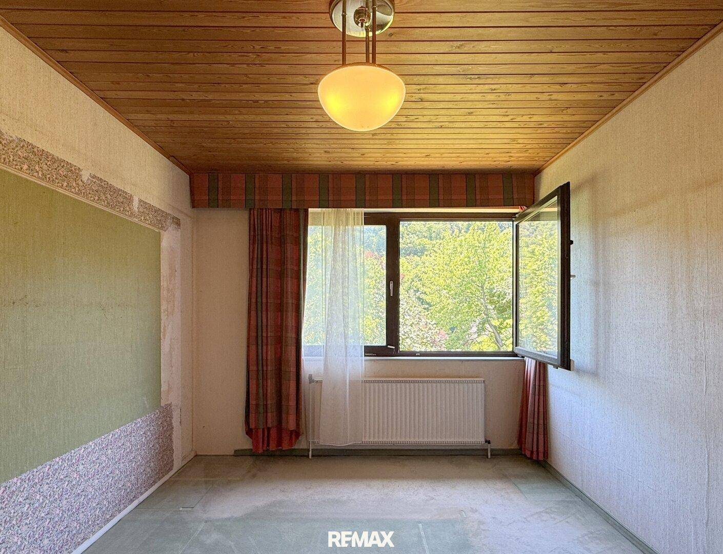 Helles Zimmer mit Holzdecke, Teppichboden und großem Fenster mit Ausblick.