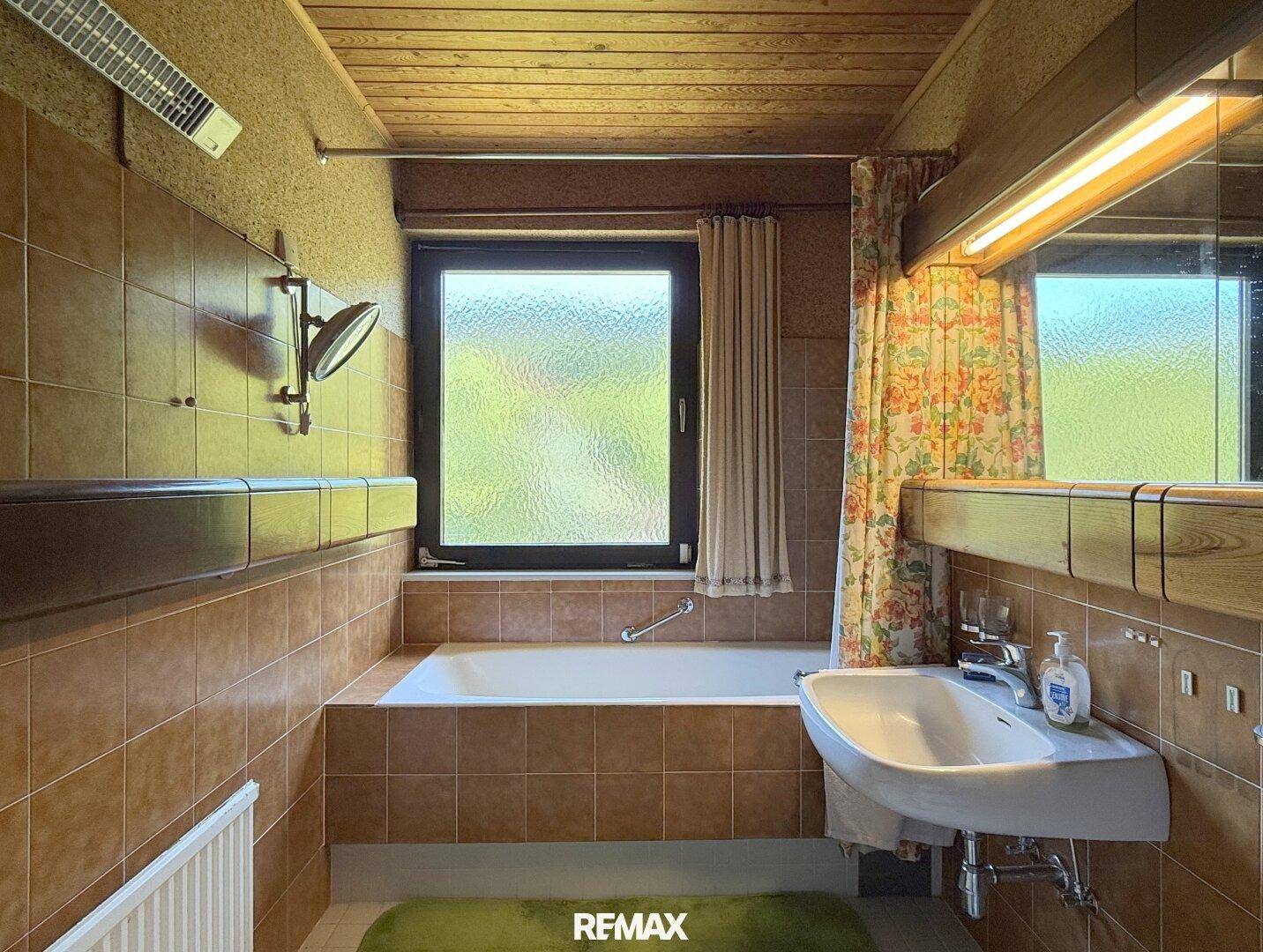 Badezimmer mit Badewanne, braunen Fliesen und Fenster mit Blick ins Grüne.