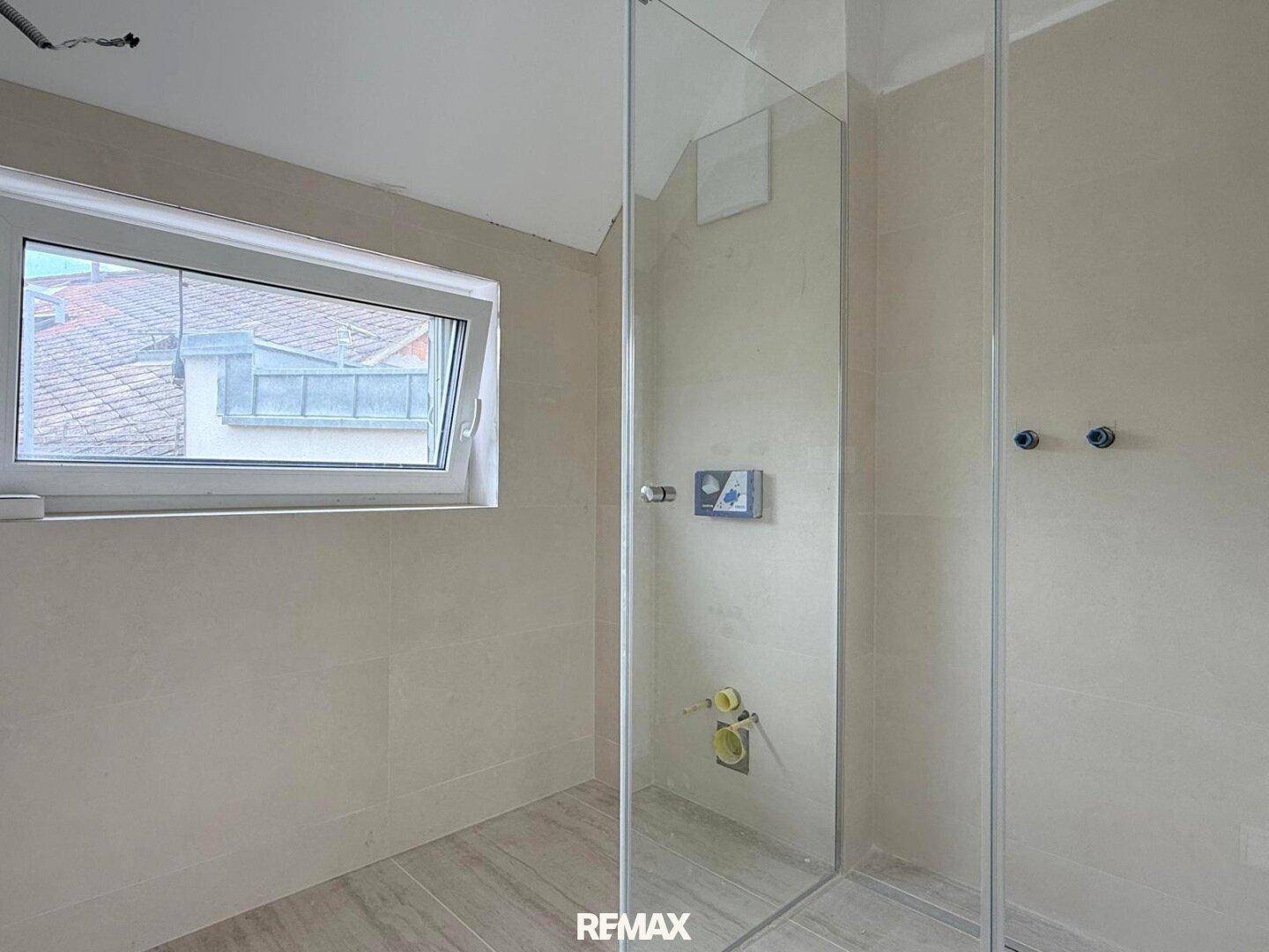 Modernes Badezimmer im Rohbau mit Duschbereich und Fenster, hell gefliest.
