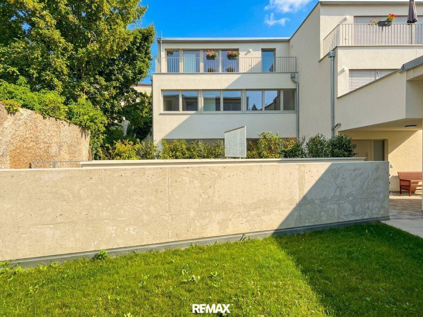Rückseite des modernen Gebäudes mit kleinem Garten und Terrasse, umgeben von Grün.