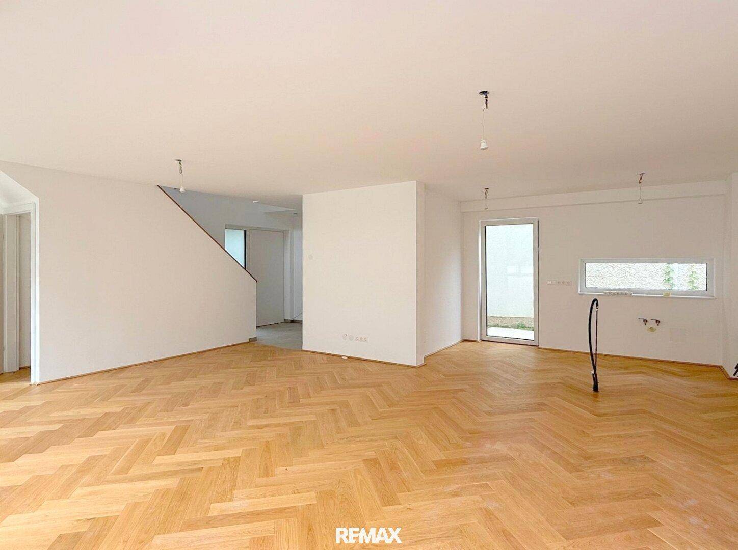 Geräumiges, leeres Wohnzimmer mit Fischgrätparkett und viel Tageslicht durch große Fenster.