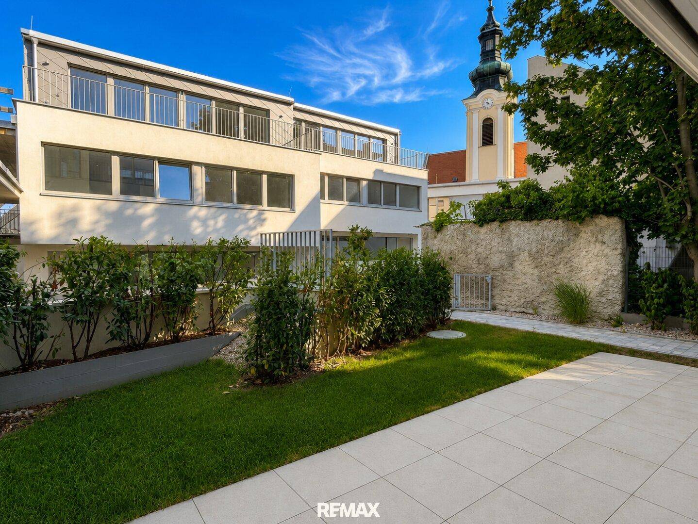 Gepflegter Gartenbereich mit Terrasse und Blick auf die moderne Wohnanlage und einen Kirchturm.