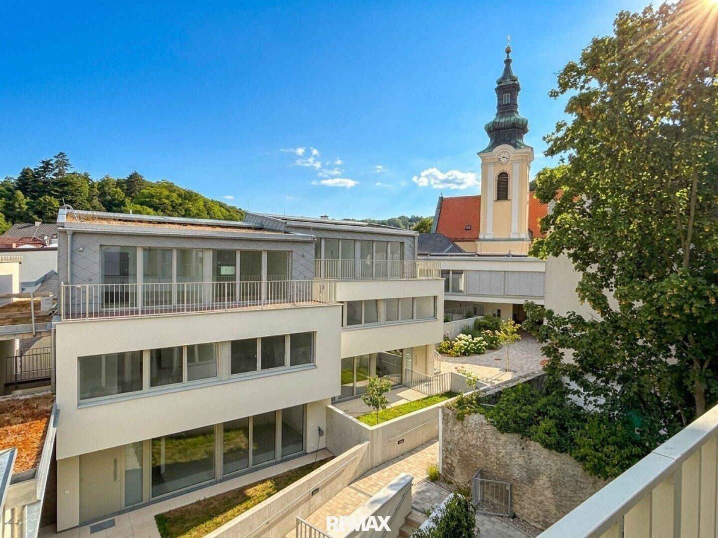 Moderne Wohnanlage mit Terrassen und Balkonen, umgeben von Grün, mit einem Kirchturm im Hintergrund.