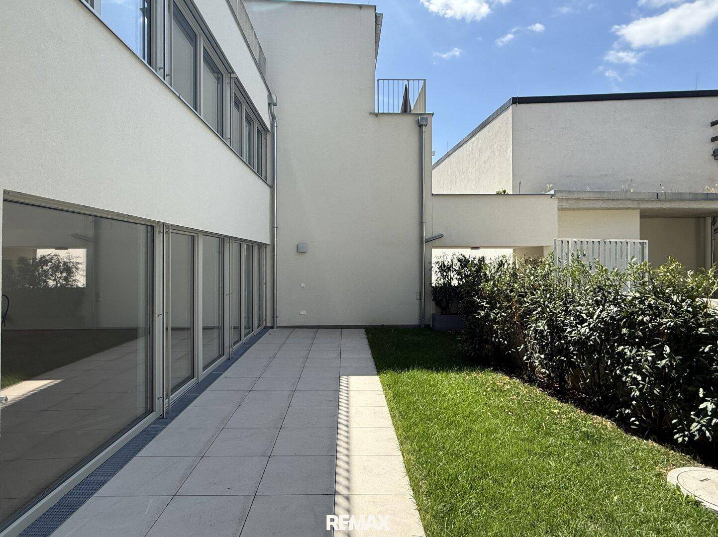 Geräumige Terrasse mit Fliesenboden und kleinem Rasenstück, umgeben von Hecken und dem Gebäude.