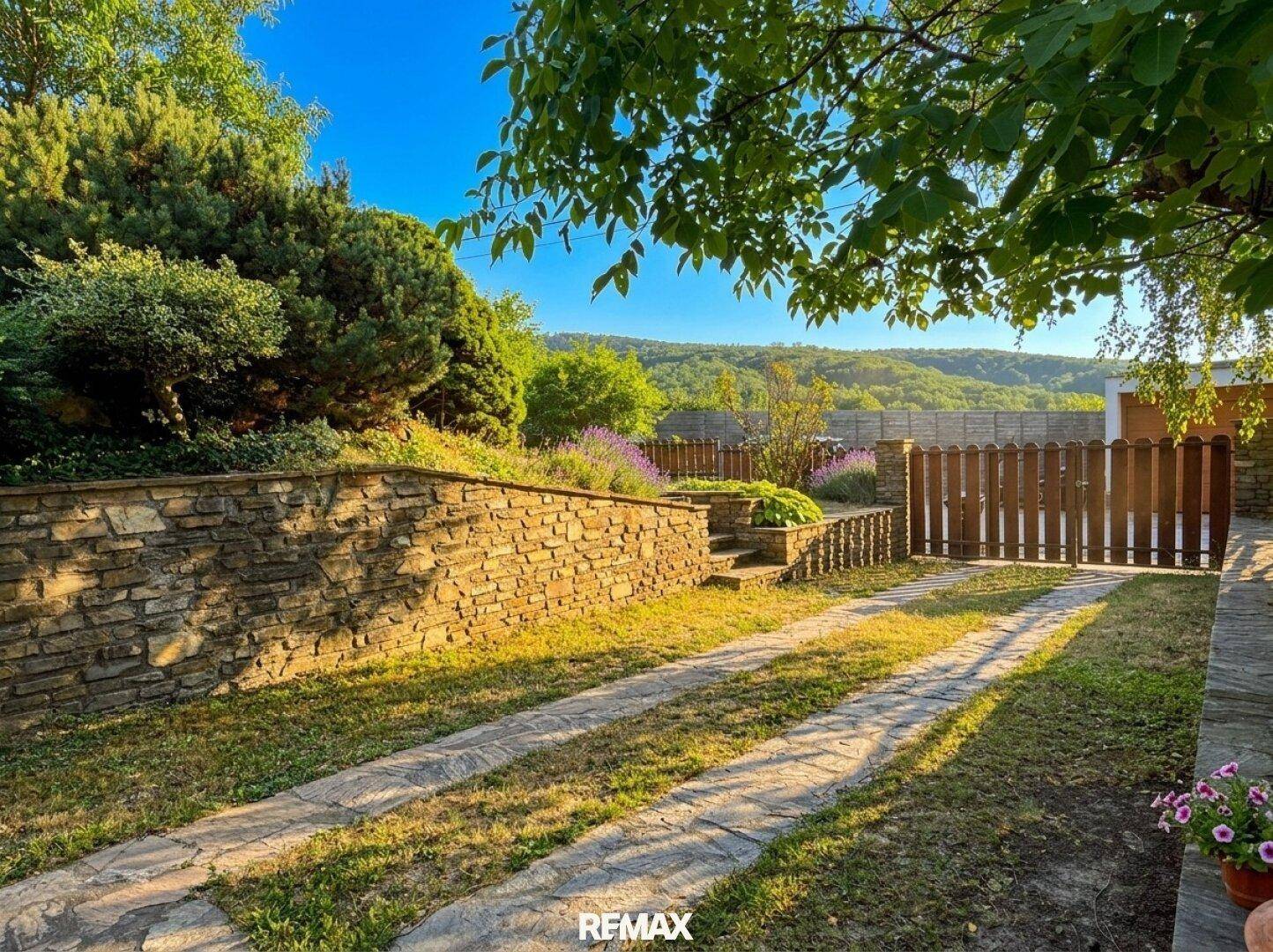 Gepflegter Gartenbereich mit Steinmauer, Zufahrt und Blick auf die umliegende Hügellandschaft.