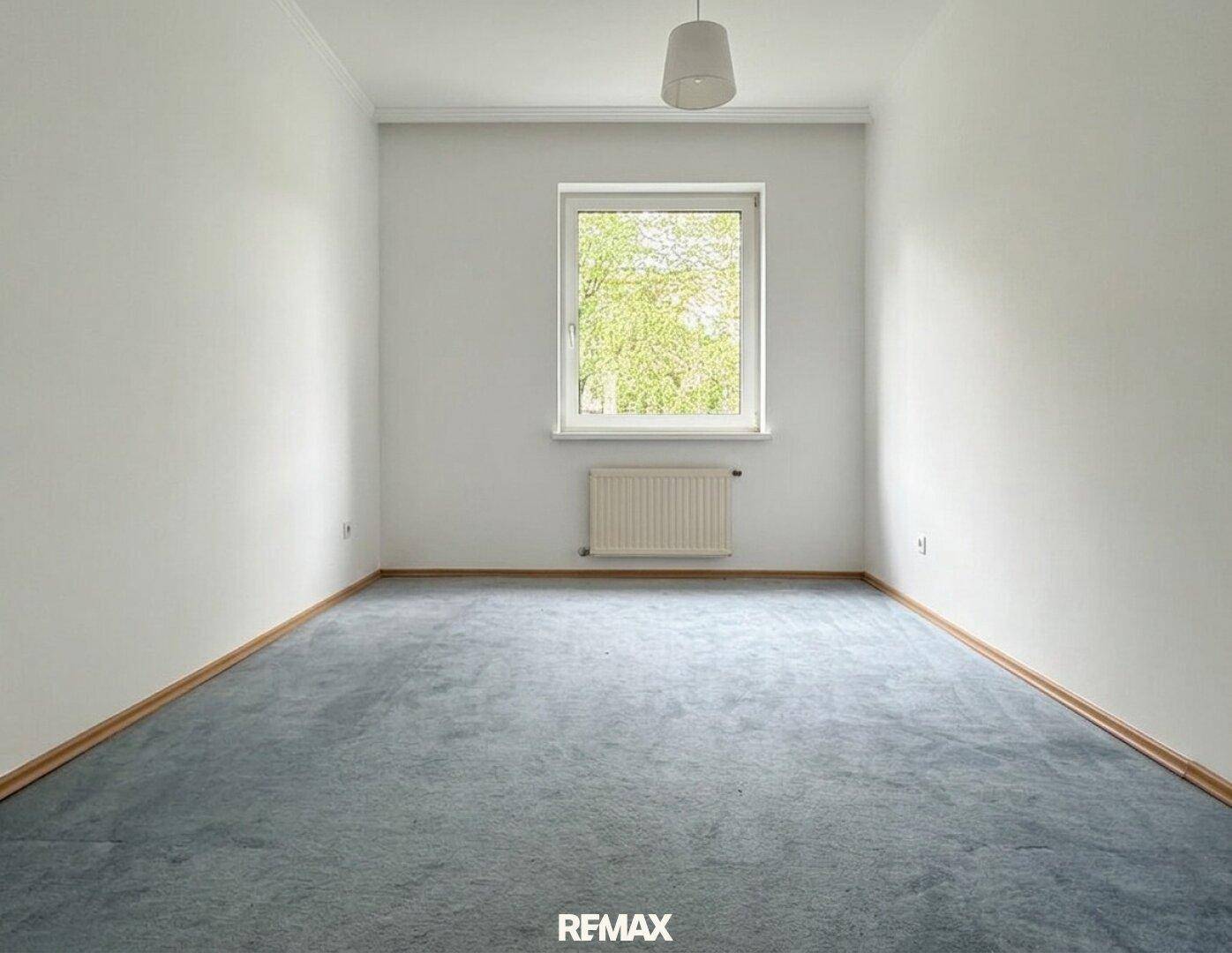 Gemütliches Schlafzimmer mit blauem Teppichboden und einem Fenster mit Blick ins Grüne.