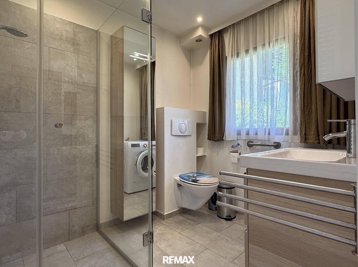 Modernes Badezimmer mit begehbarer Dusche, WC und Waschmaschine, hell gefliest.