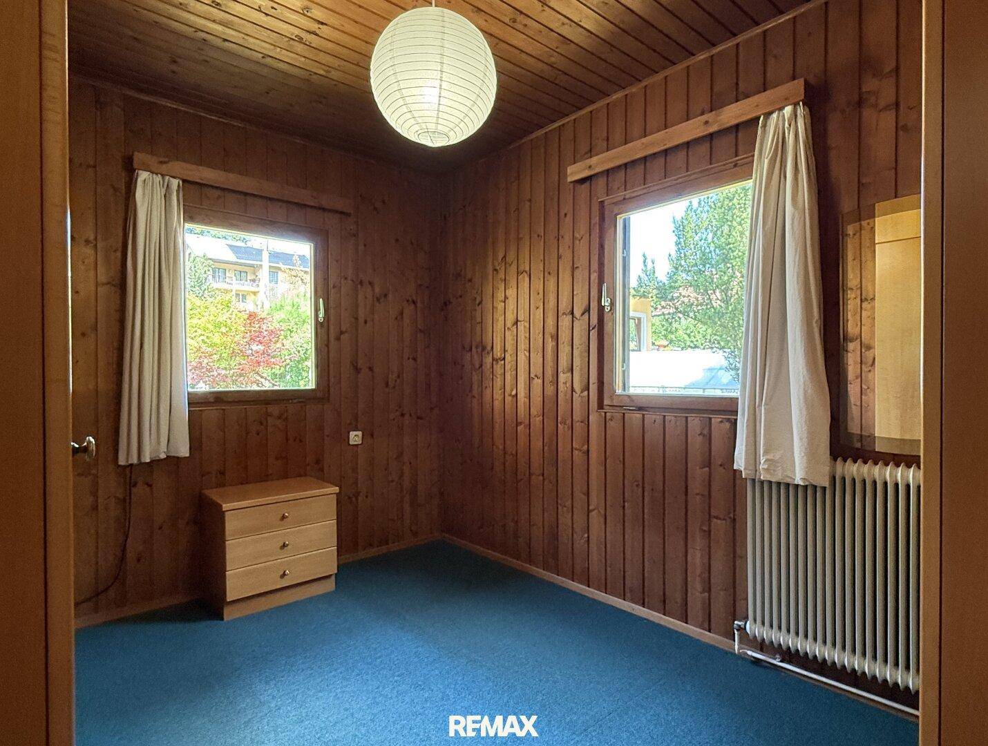 Schlafzimmer mit Holzwänden, Teppichboden und zwei Fenstern mit Blick ins Grüne.
