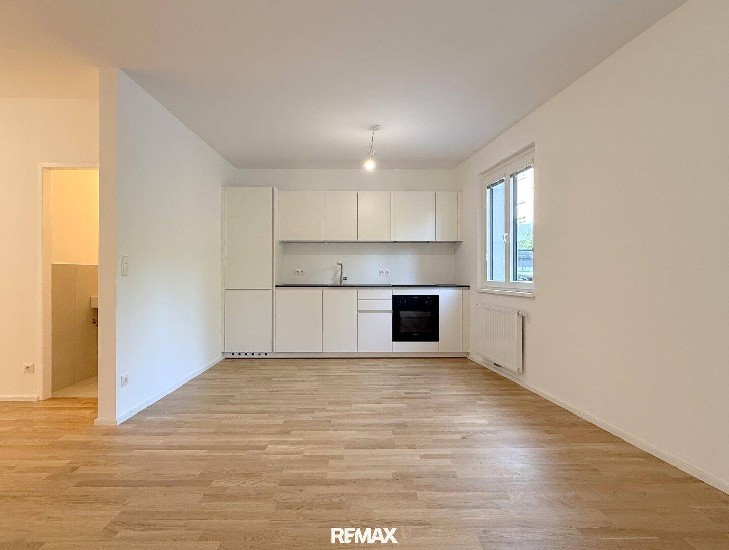 Heller Wohnbereich mit moderner weißer Einbauküche, Fenster und Holzboden.