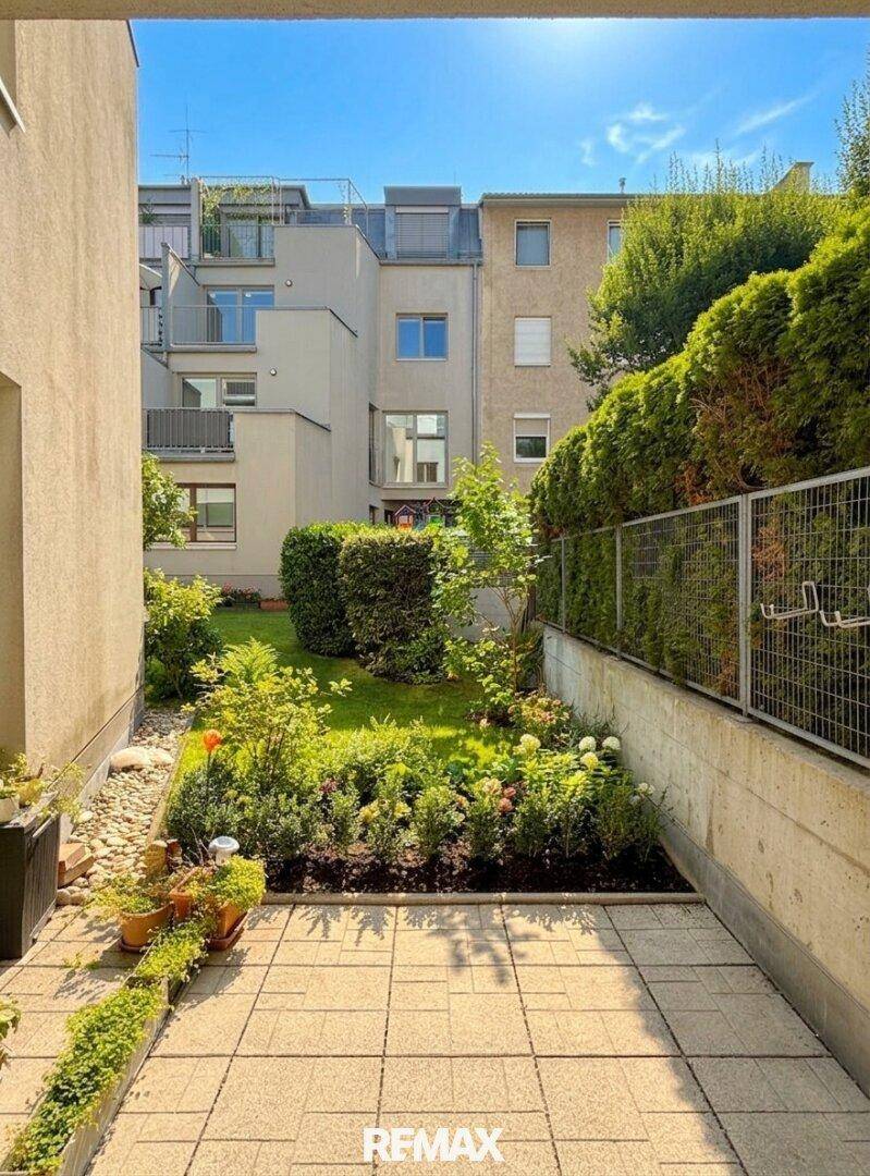 Sonniger Gartenbereich mit gepflasterter Terrasse, Blumenbeeten und Blick auf umliegende Gebäude.