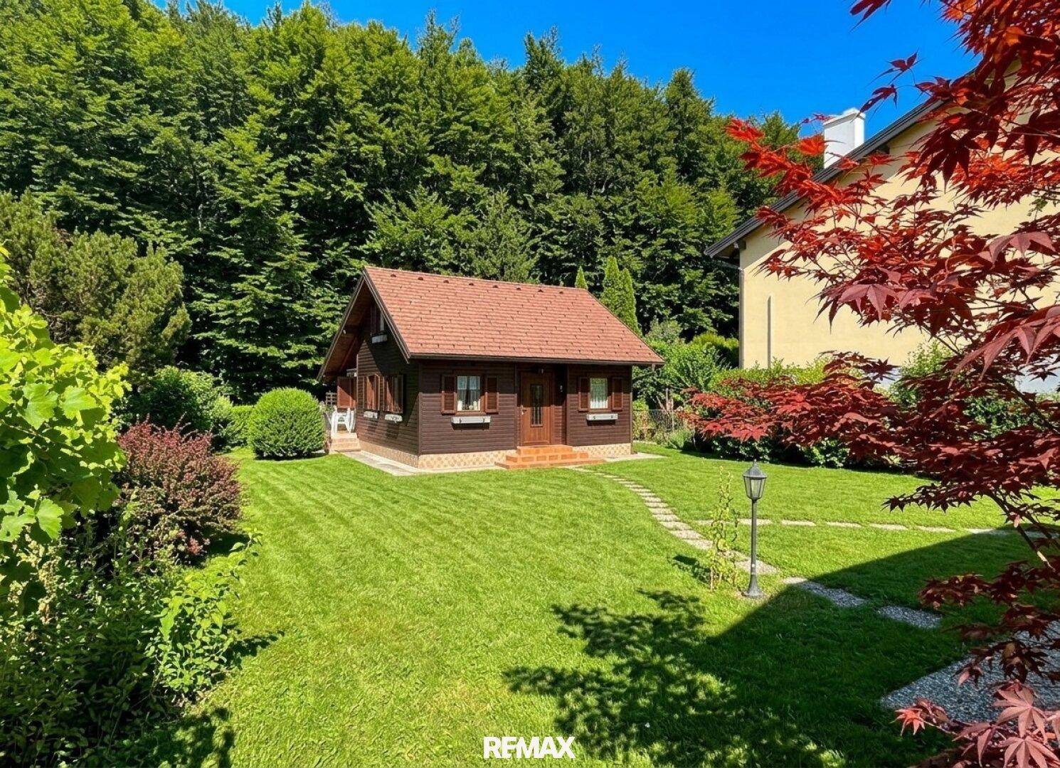 Panoramablick auf das Holzhaus und den weitläufigen, sonnigen Garten mit Bäumen.