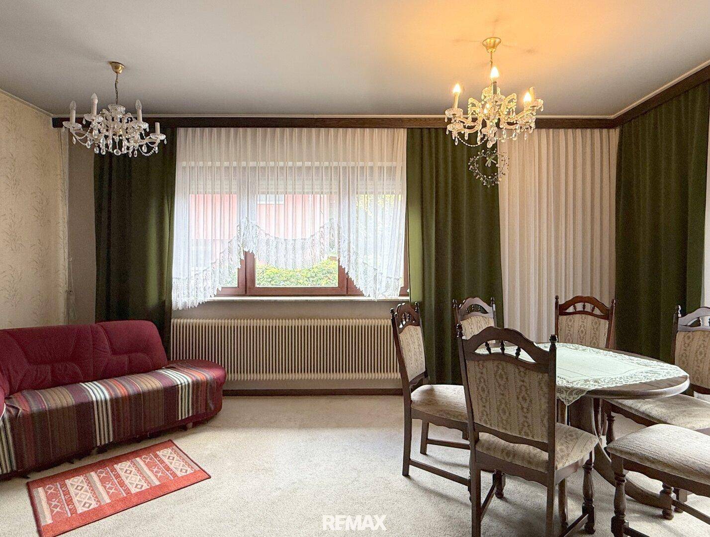 Gemütlicher Wohn- und Essbereich mit Teppichboden, rotem Sofa und klassischem Kronleuchter.