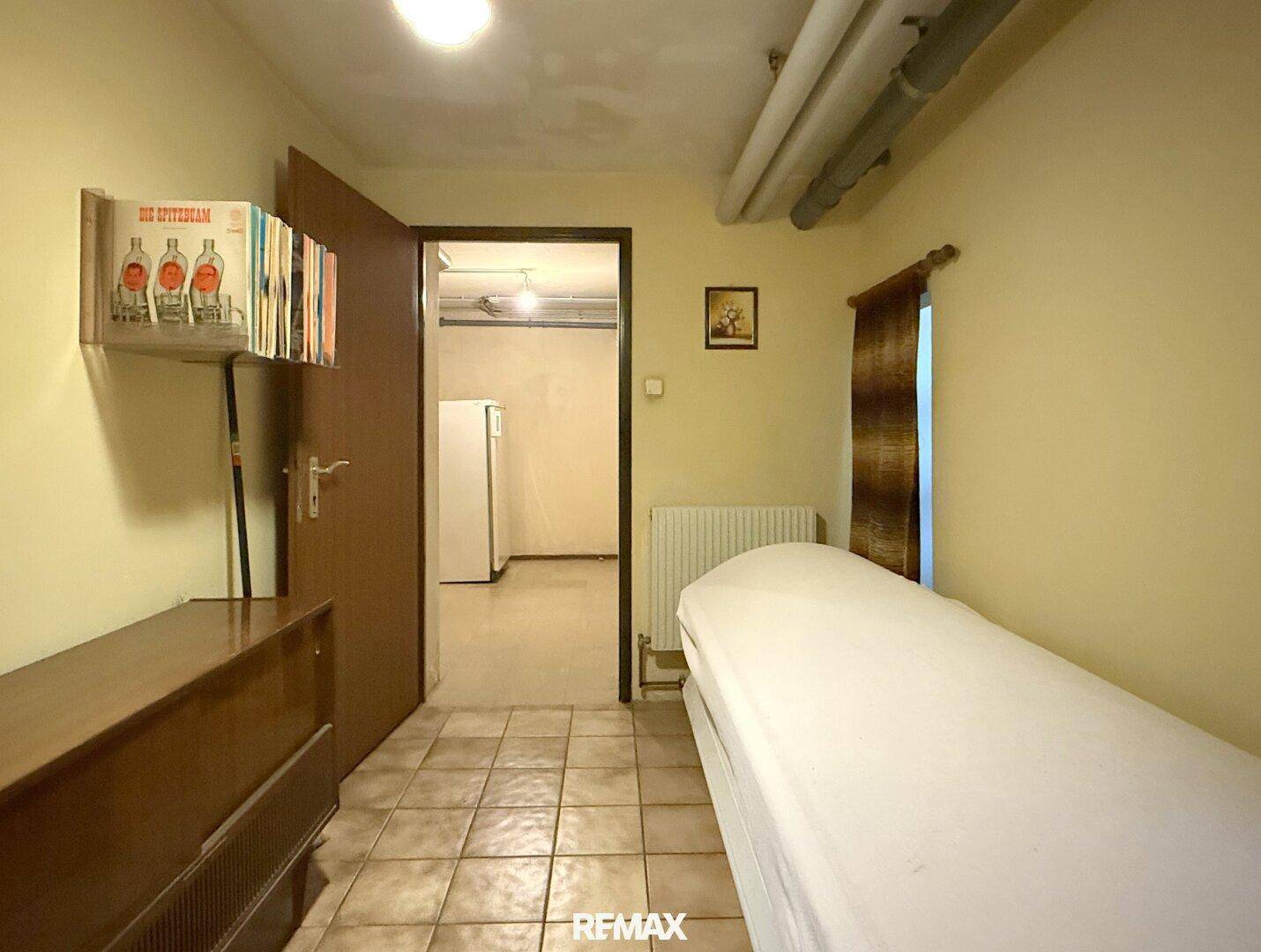 Kellerraum mit Fliesenboden, Bett und Zugang zu weiteren Kellerräumen.