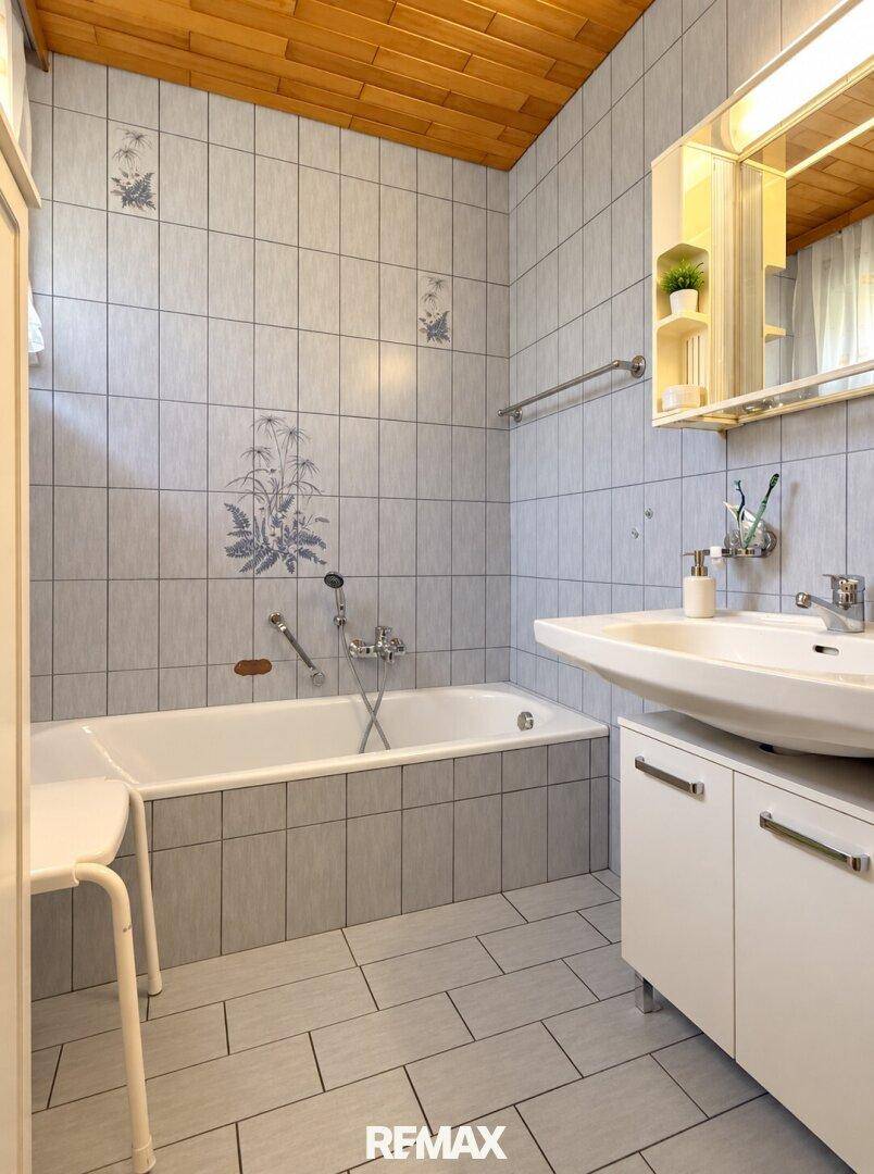 Badezimmer mit Badewanne, Waschbecken und Holzdecke, ausgestattet mit hellgrauen Fliesen.