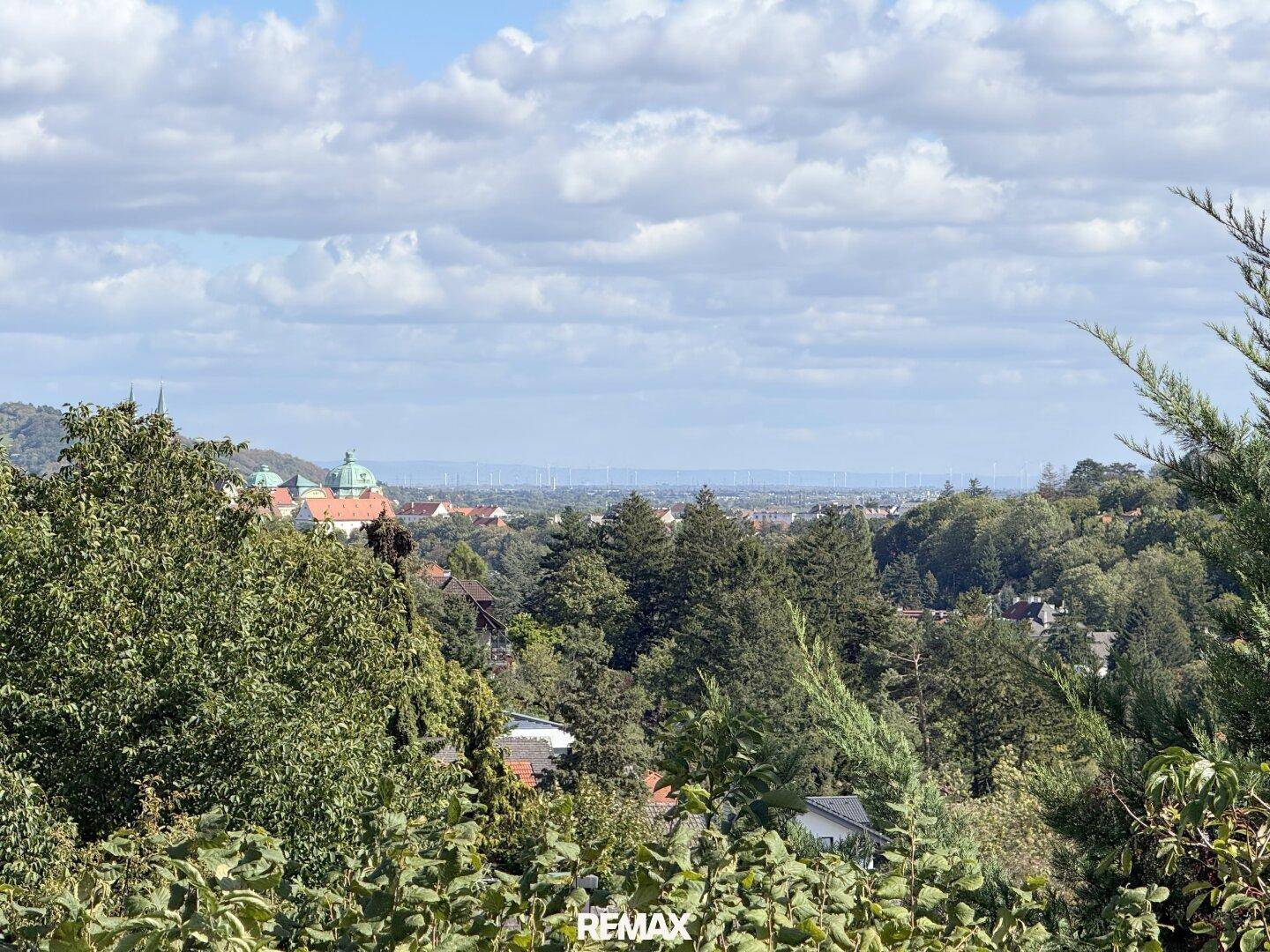 Weitläufiger Blick über die grüne Landschaft mit entfernten Gebäuden und Windrädern am Horizont.