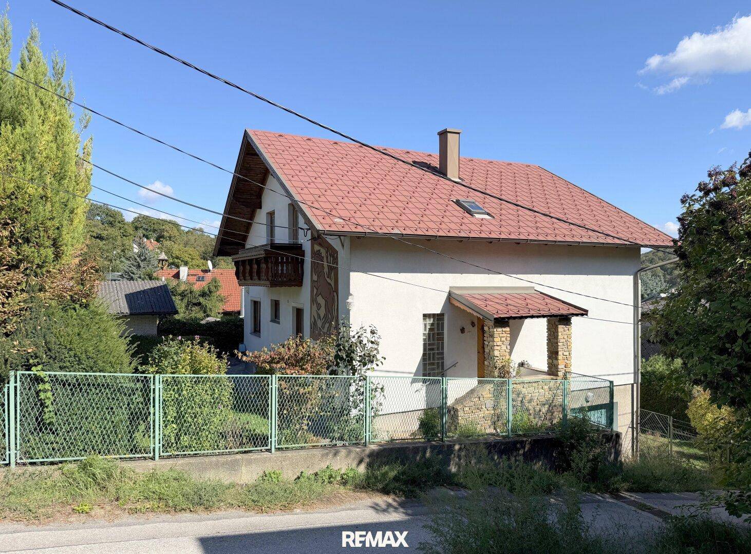 Gesamtansicht des Hauses von der Straße aus mit rotem Ziegeldach und umzäuntem Grundstück.