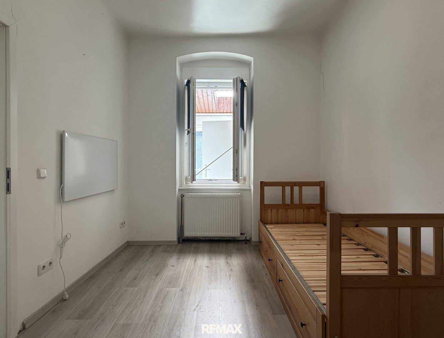 Schlafzimmer mit Einzelbett aus Holz, Laminatboden und Fenster.