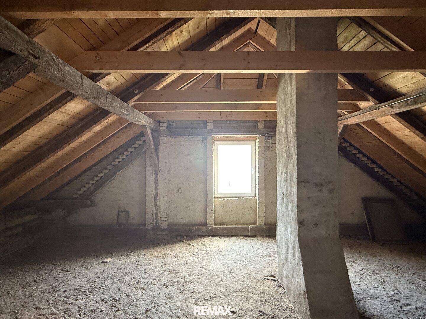 Unausgebauter Dachboden mit sichtbaren Holzbalken und kleinem Fenster.