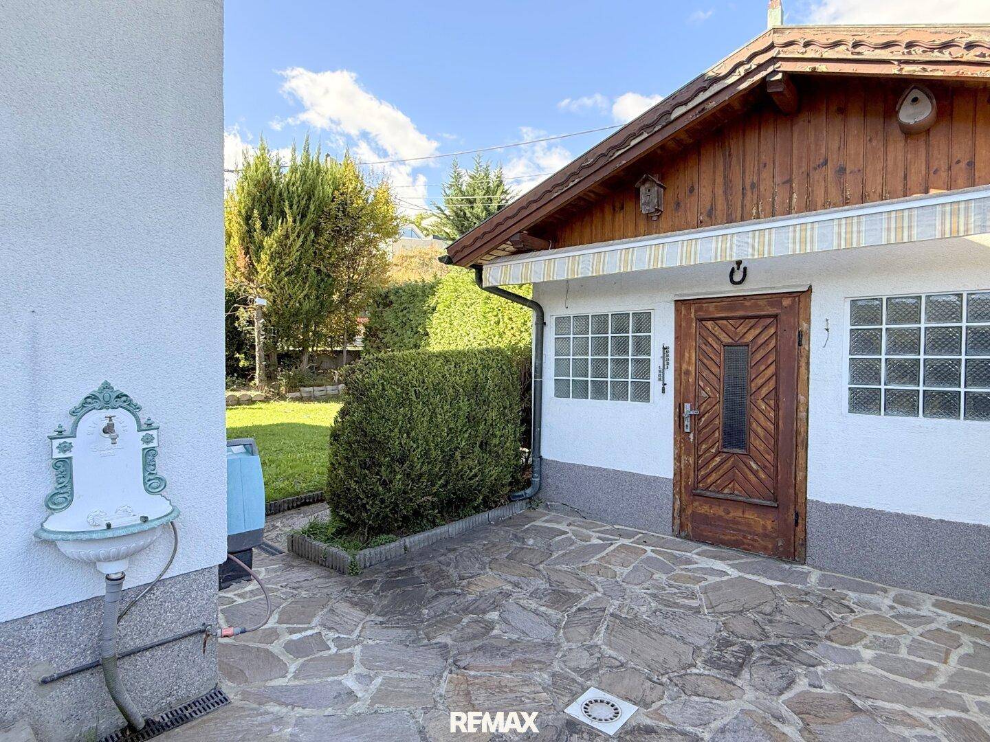 Gepflasterter Terrassenbereich neben einem Nebengebäude mit dekorativem Wasserhahn.