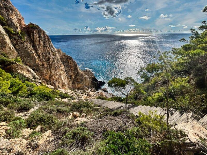 Panoramablick von einem Klippenpfad auf das glitzernde Meer und die felsige Küste mit Holztreppen.
