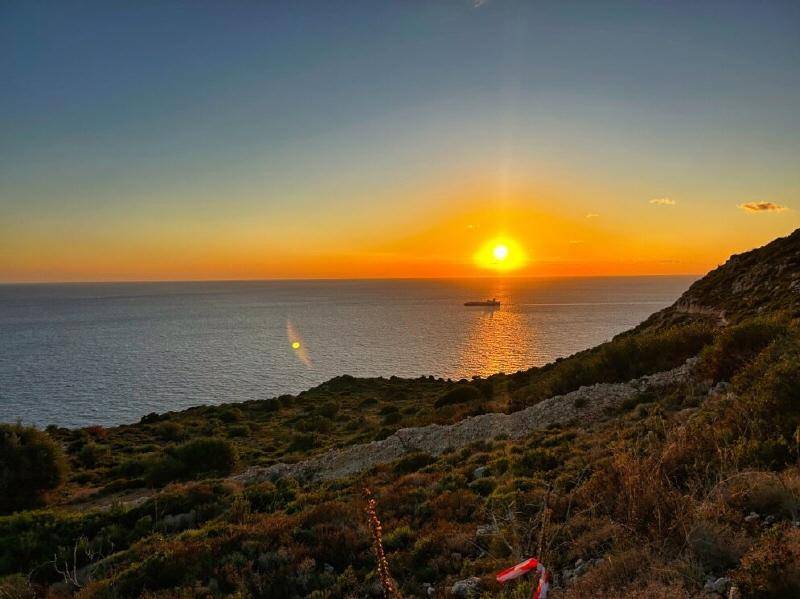 Spektakulärer Sonnenuntergang über dem Meer, gesehen von einem Hügel mit spärlicher Vegetation und einem Schiff am Horizont.