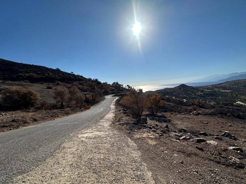 Panoramablick auf eine kurvenreiche Straße durch trockene Landschaft mit Meer- und Bergblick bei hellem Sonnenschein.