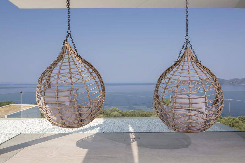 Zwei stilvolle Hängesessel auf einem Balkon mit Glasgeländer und weitem Blick über das Meer.