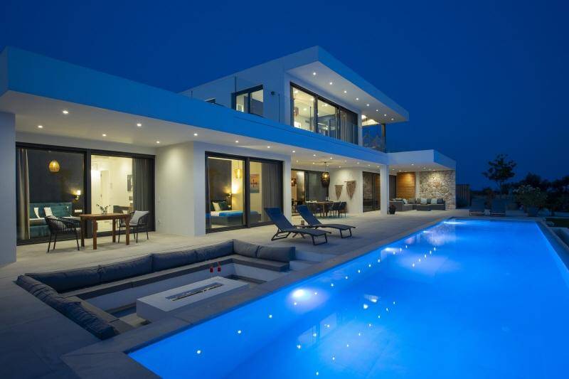 Beleuchtete moderne Villa bei Nacht mit leuchtendem Pool und gemütlicher versunkener Sitzgelegenheit mit Feuerstelle.