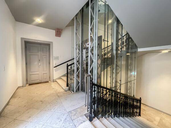 Moderner Glasaufzug neben einer historischen Steintreppe mit schmiedeeisernem Geländer in einem Treppenhaus.