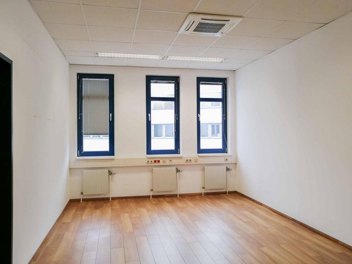 Leeres Büro mit Holzboden, weißen Wänden und drei Fenstern mit Heizkörpern darunter.