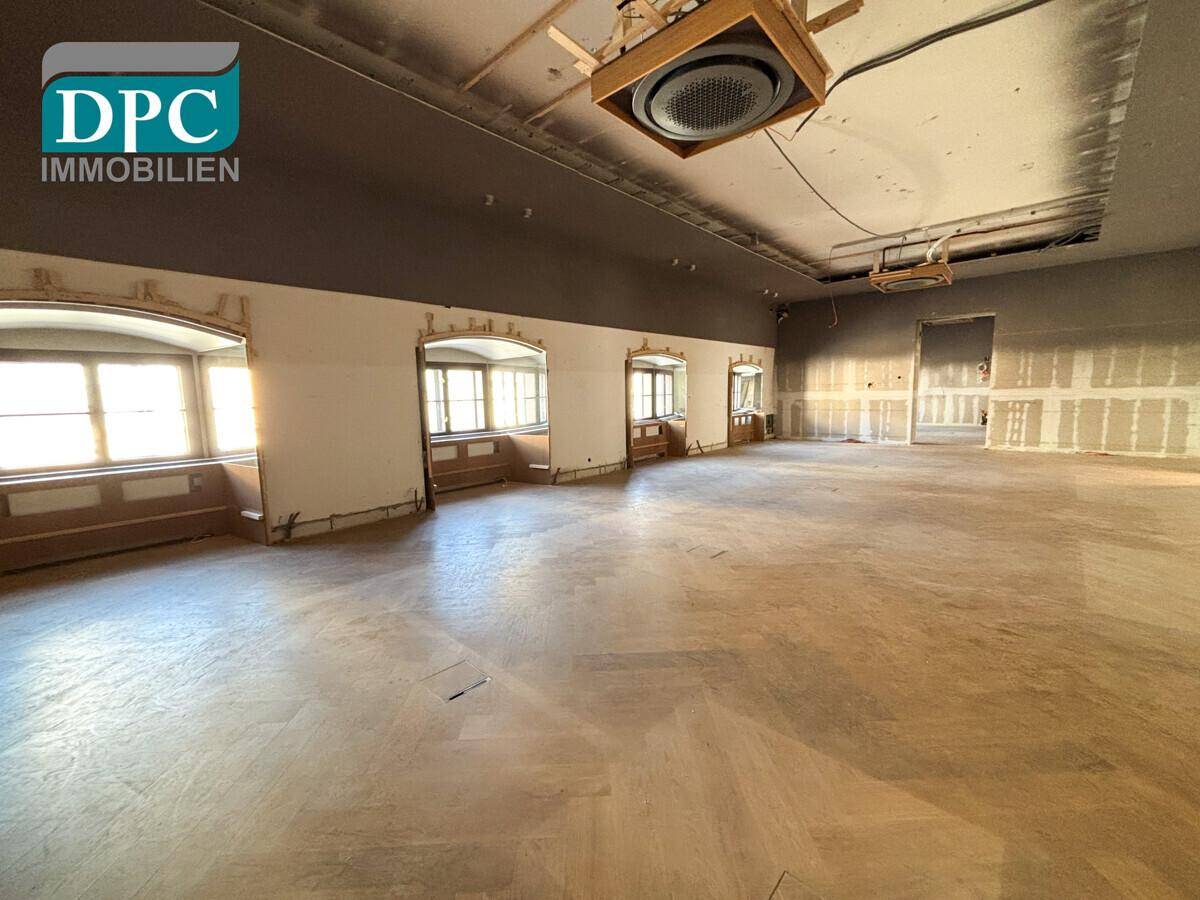 Renovierungsbedürftiger Innenraum mit Fischgrätparkett, sichtbaren Installationen und hohen Fenstern.