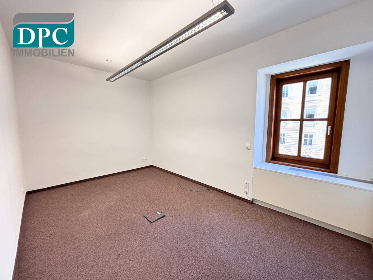 Kleinerer Büroraum mit Teppichboden und einem Fenster, das Tageslicht spendet.