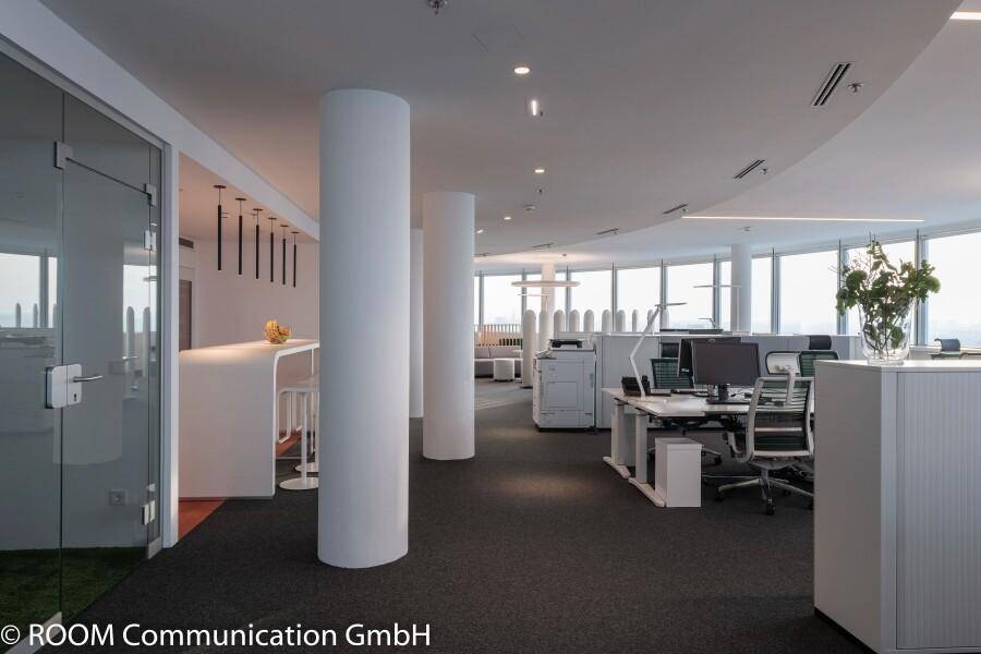 Geräumiger Großraumbüro mit weißen Säulen, modernen Arbeitsplätzen und viel Tageslicht durch große Fenster.