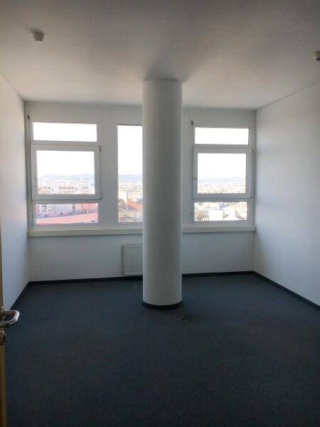 Heller Büroraum mit einer zentralen Säule, großen Fenstern und einem dunklen Teppichboden, der viel Tageslicht hereinlässt.