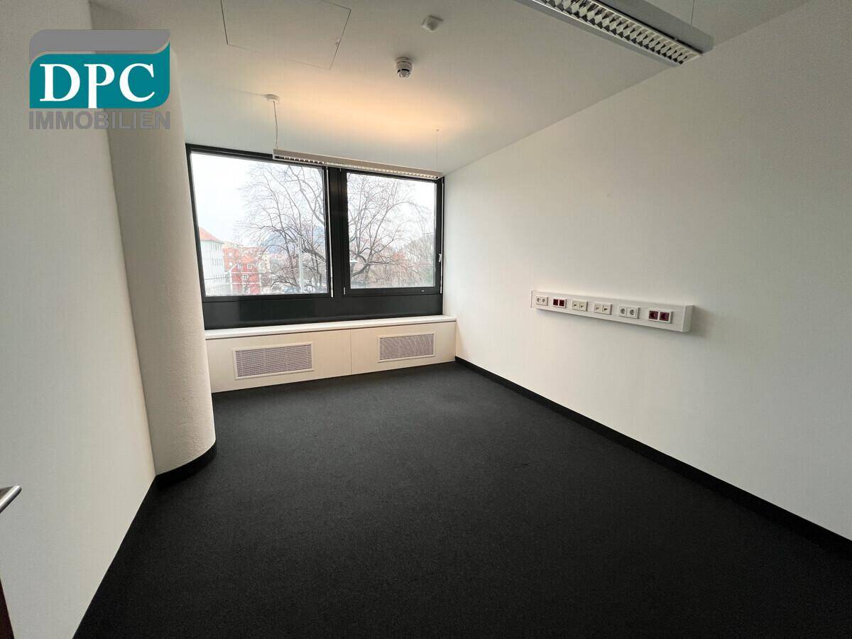 Geräumiges Büro mit großen Fenstern, schwarzem Teppichboden und viel natürlichem Licht.