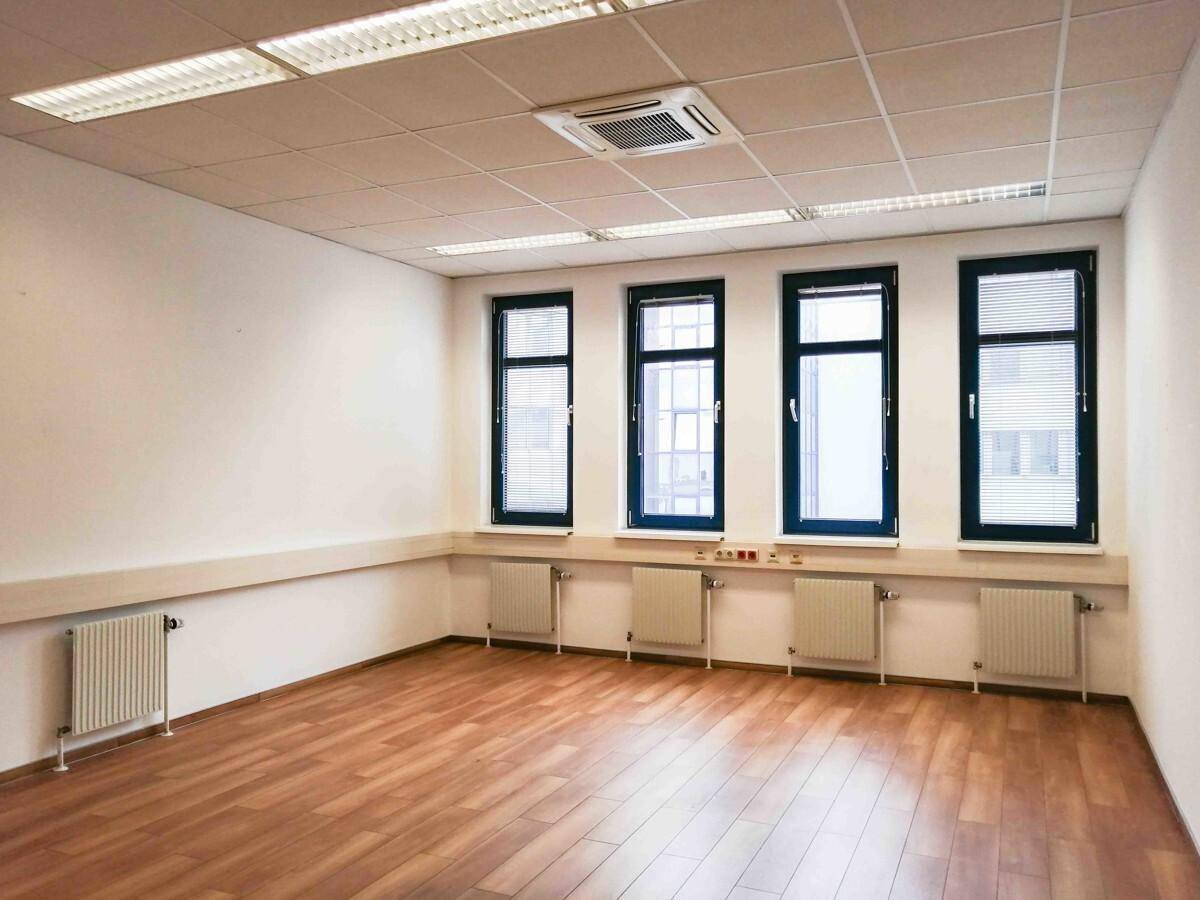 Leeres Büro mit Holzboden, weißen Wänden und vier Fenstern mit Heizkörpern darunter.