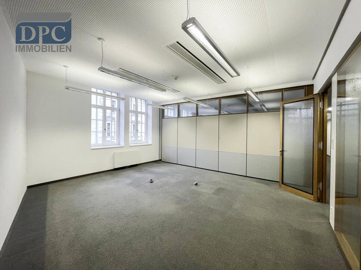 Leerer Büroraum mit grauem Teppichboden, weißen Wänden, Fenstern und einer Trennwand für flexible Gestaltung.