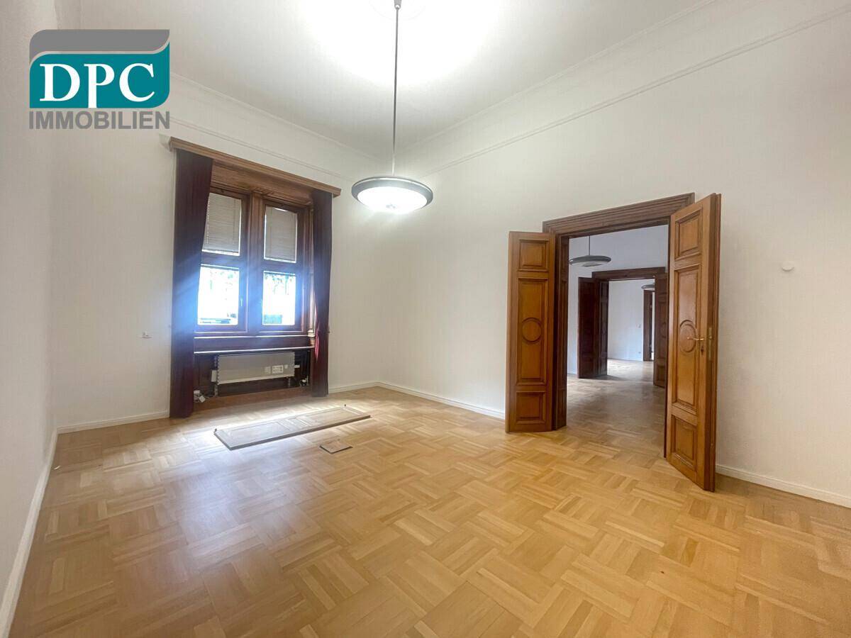 Geräumiger Raum mit Fischgrätparkett, hohen Decken und Blick in angrenzende Zimmer durch offene Holztüren.