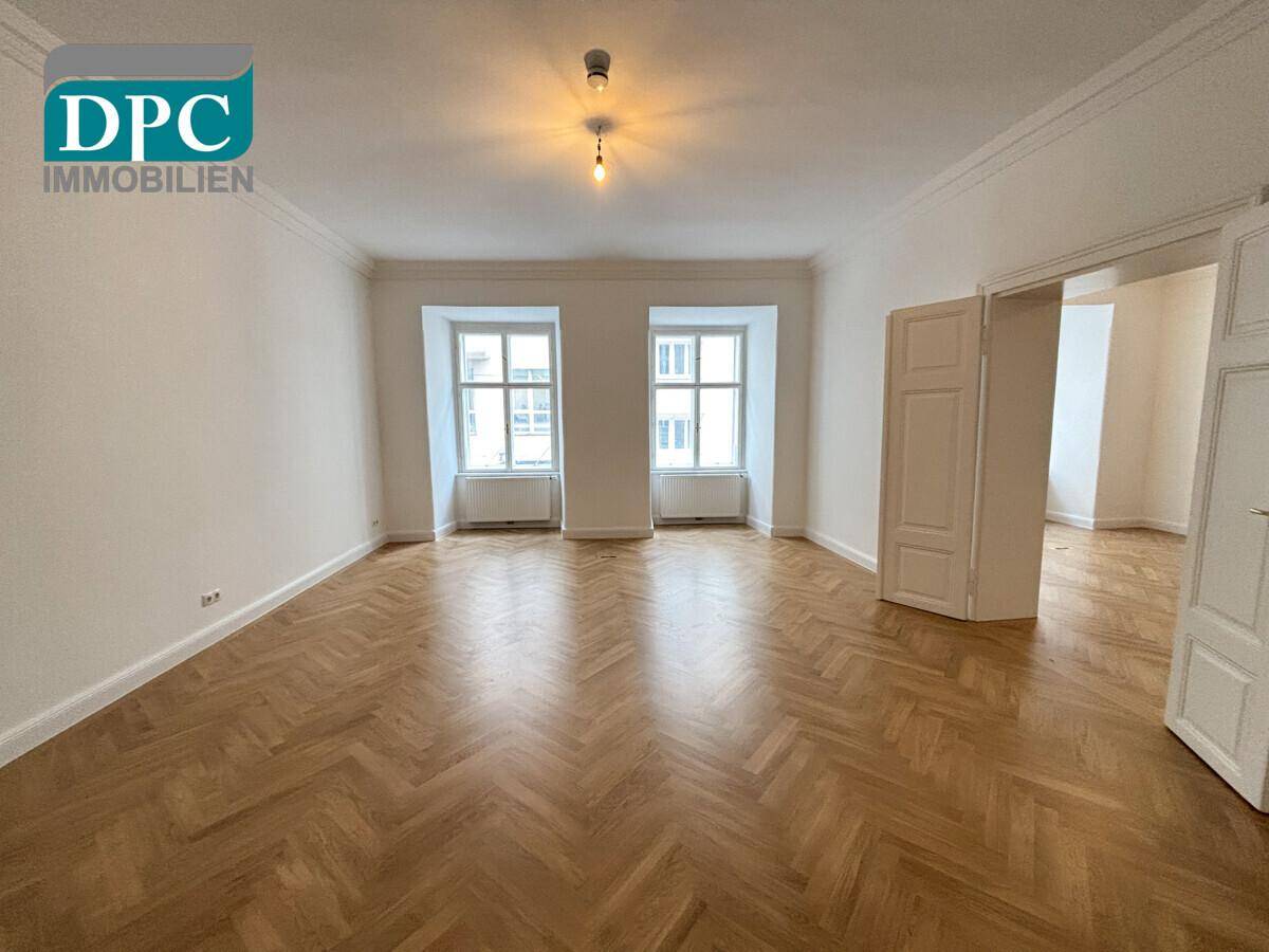 Geräumiges Zimmer mit zwei großen Fenstern, Fischgrätparkett und hohen Decken, ideal für Wohnzwecke.