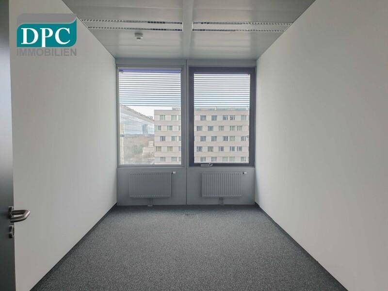 Kleines Büro mit Teppichboden, zwei Fenstern und Blick auf andere Gebäude.