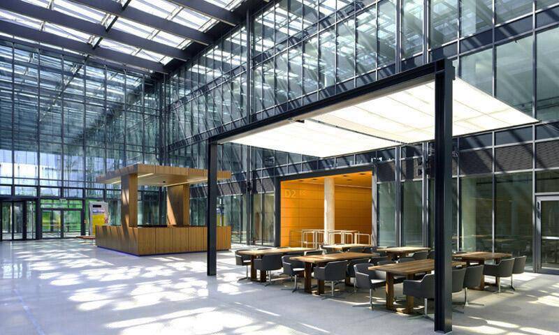 Geräumige Lobby mit Glasdach, Sitzgelegenheiten und einem Empfangsbereich in einem modernen Bürogebäude.