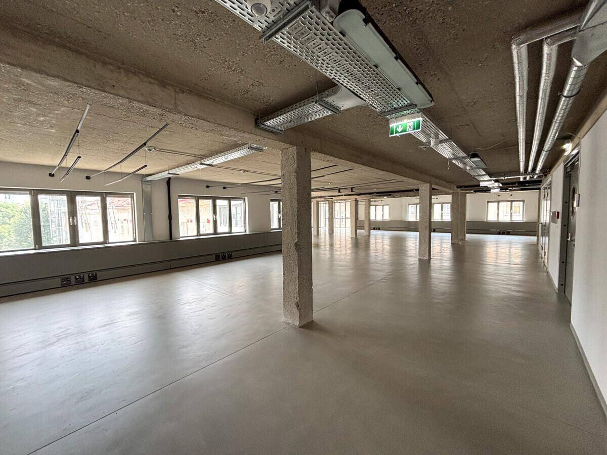Großer, offener Büroraum mit Betonboden, sichtbaren Deckeninstallationen und vielen Fenstern für natürliches Licht.
