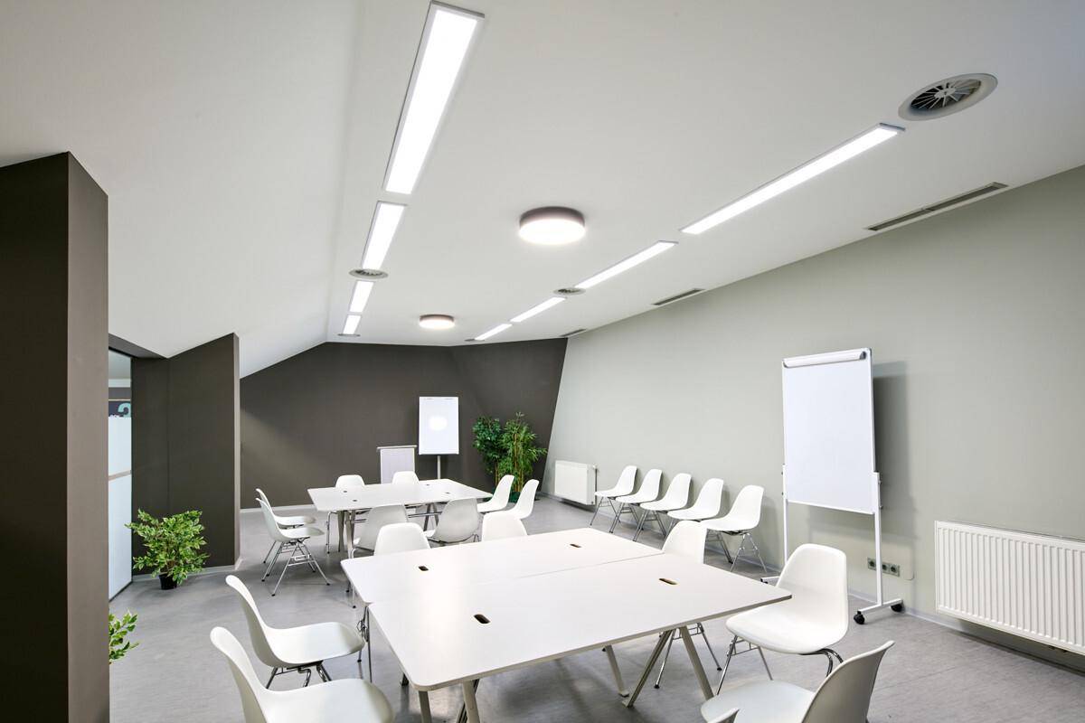 Heller Konferenzraum mit weißen Tischen und Stühlen, effizienter Beleuchtung und schrägen Decken.