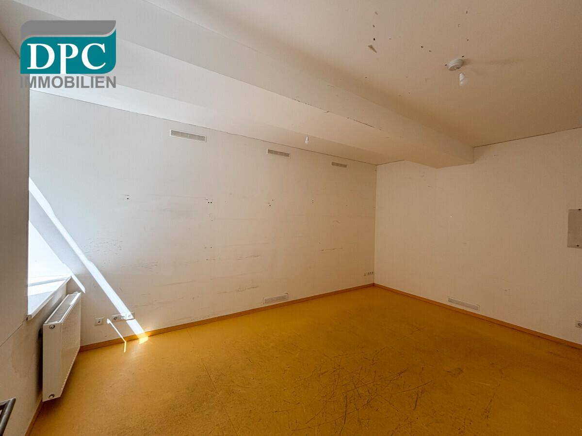 Kleiner, leerer Raum mit gelbem Boden und weiß gestrichenen Wänden, mit einem Fenster für Tageslicht.