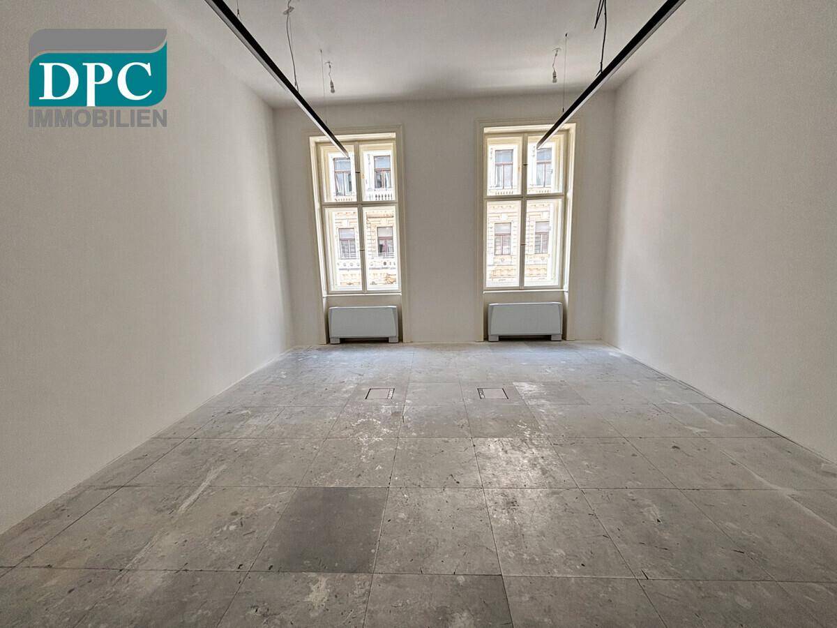 Geräumiger, heller Gewerberaum mit zwei großen Fenstern und unfertigem Betonboden, bereit für den Innenausbau.